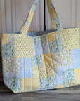A large quilted patchwork tote bag with yellow, blue, and white fabrics, standing on a gray wood floor next to a potted green plant.