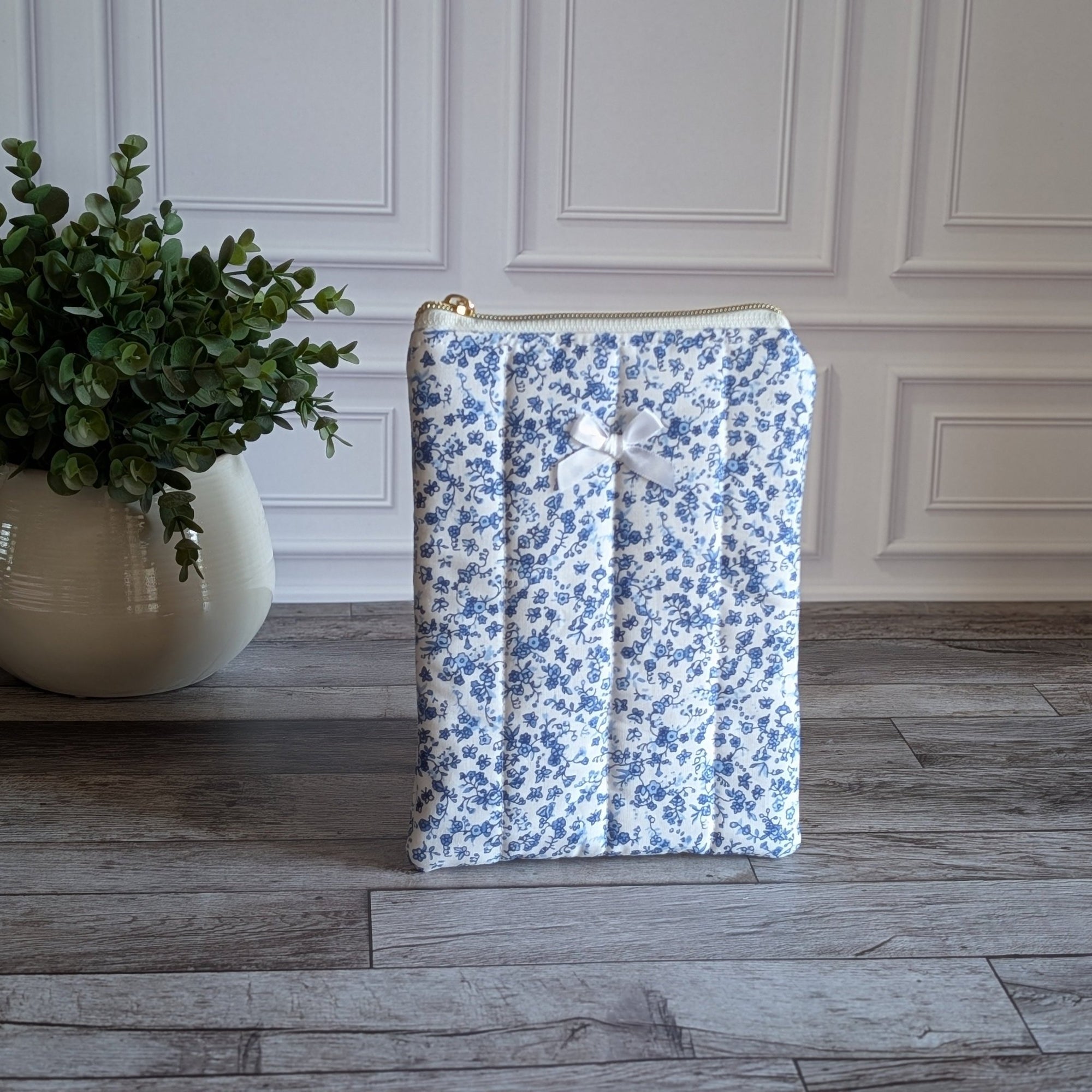A quilted fabric book or device sleeve with a blue and white floral pattern, featuring a white bow on the front and a gold zipper at the top, set on a gray wood floor next to a potted green plant.