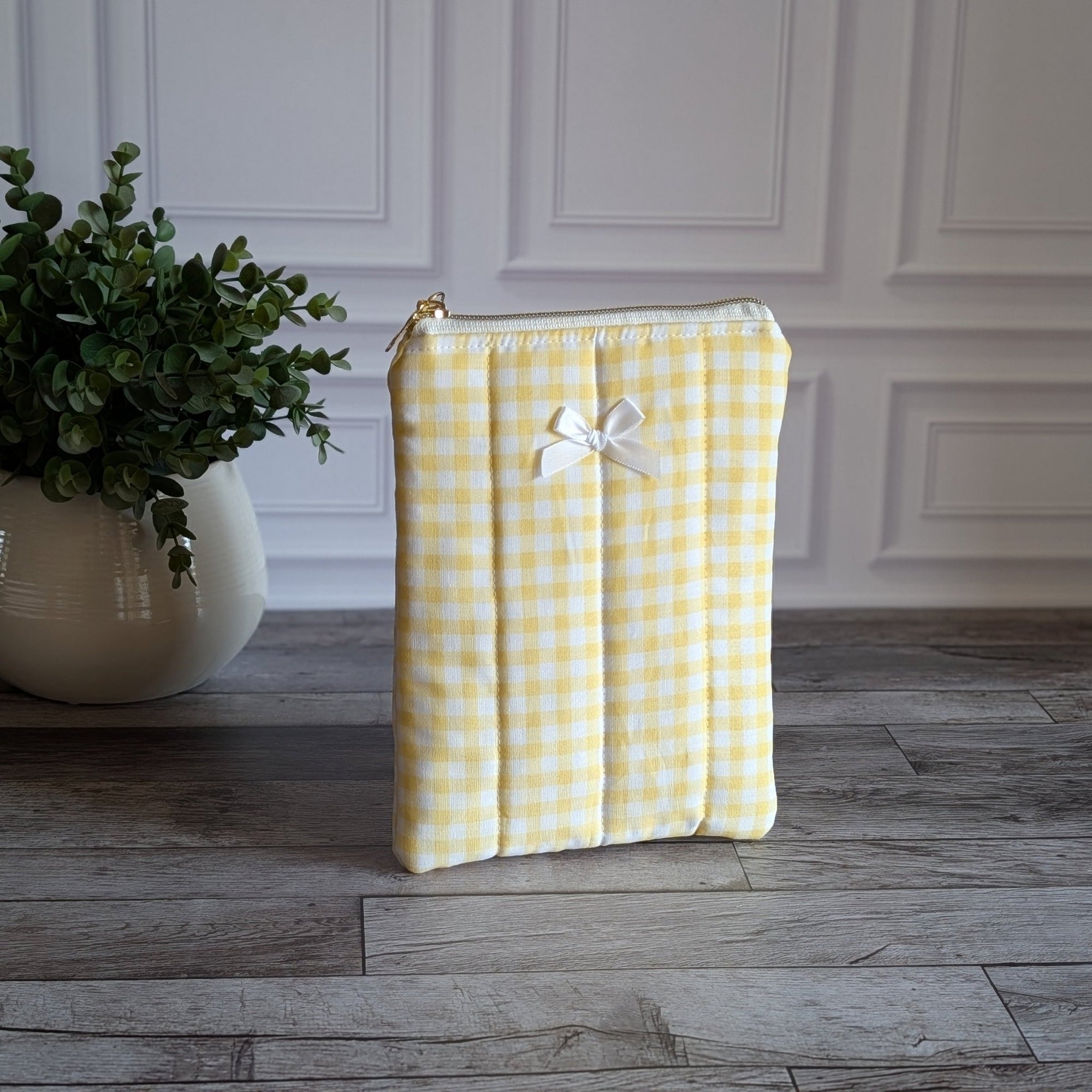 A yellow and white gingham quilted pouch with a small white bow on the front, standing on a gray wood floor in front of a white paneled wall next to a potted green plant.