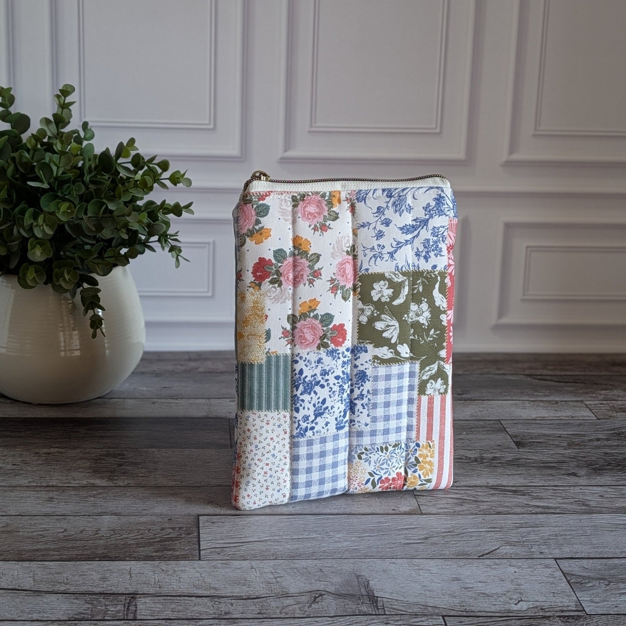 A rectangular patchwork quilted zippered pouch with a floral and gingham pattern, standing on a gray wood floor next to a potted green plant against a white paneled wall.