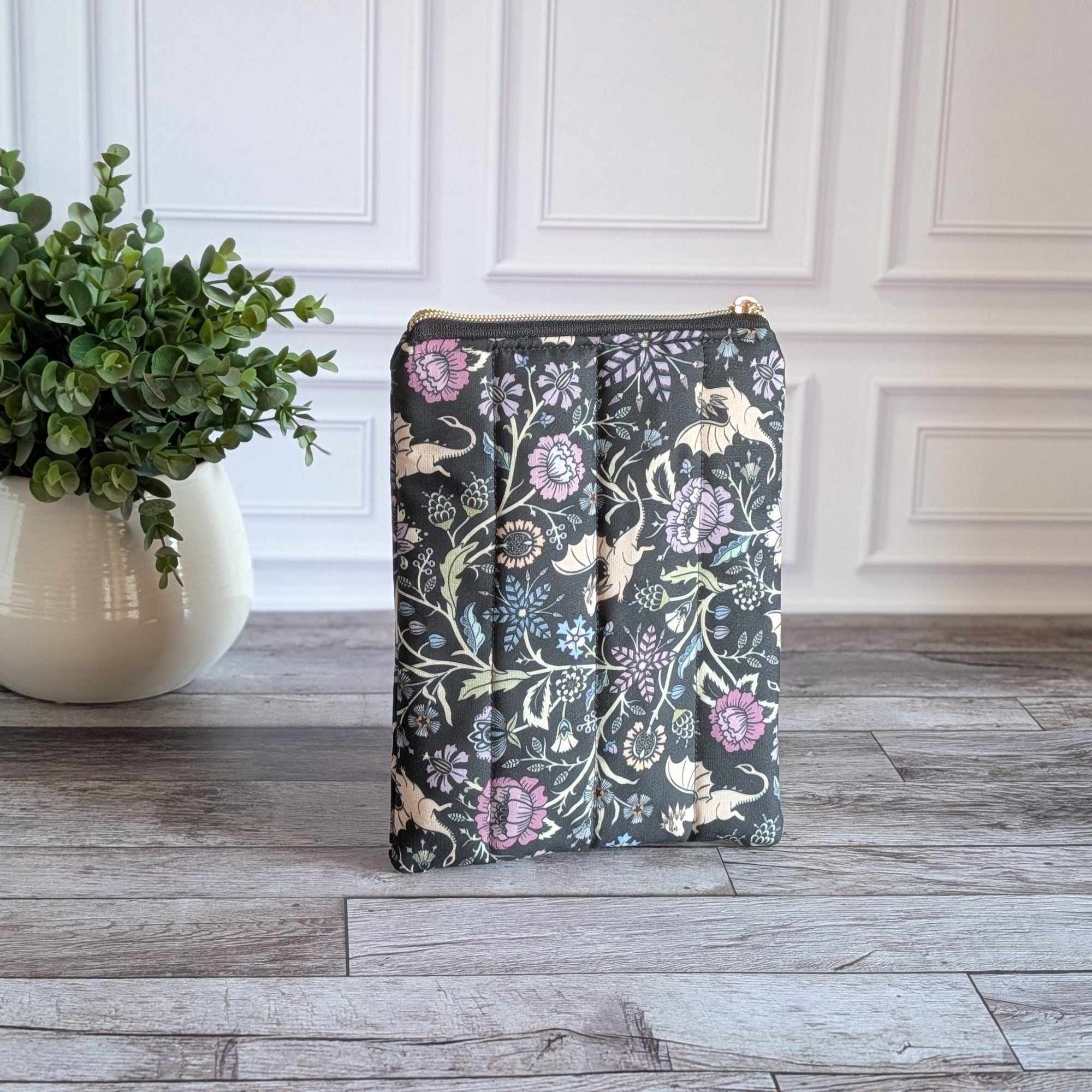 A vertical zippered pouch with a dark green, purple, and black floral pattern featuring beige dragons, standing on a gray wood table next to a potted green plant against a white paneled wall.