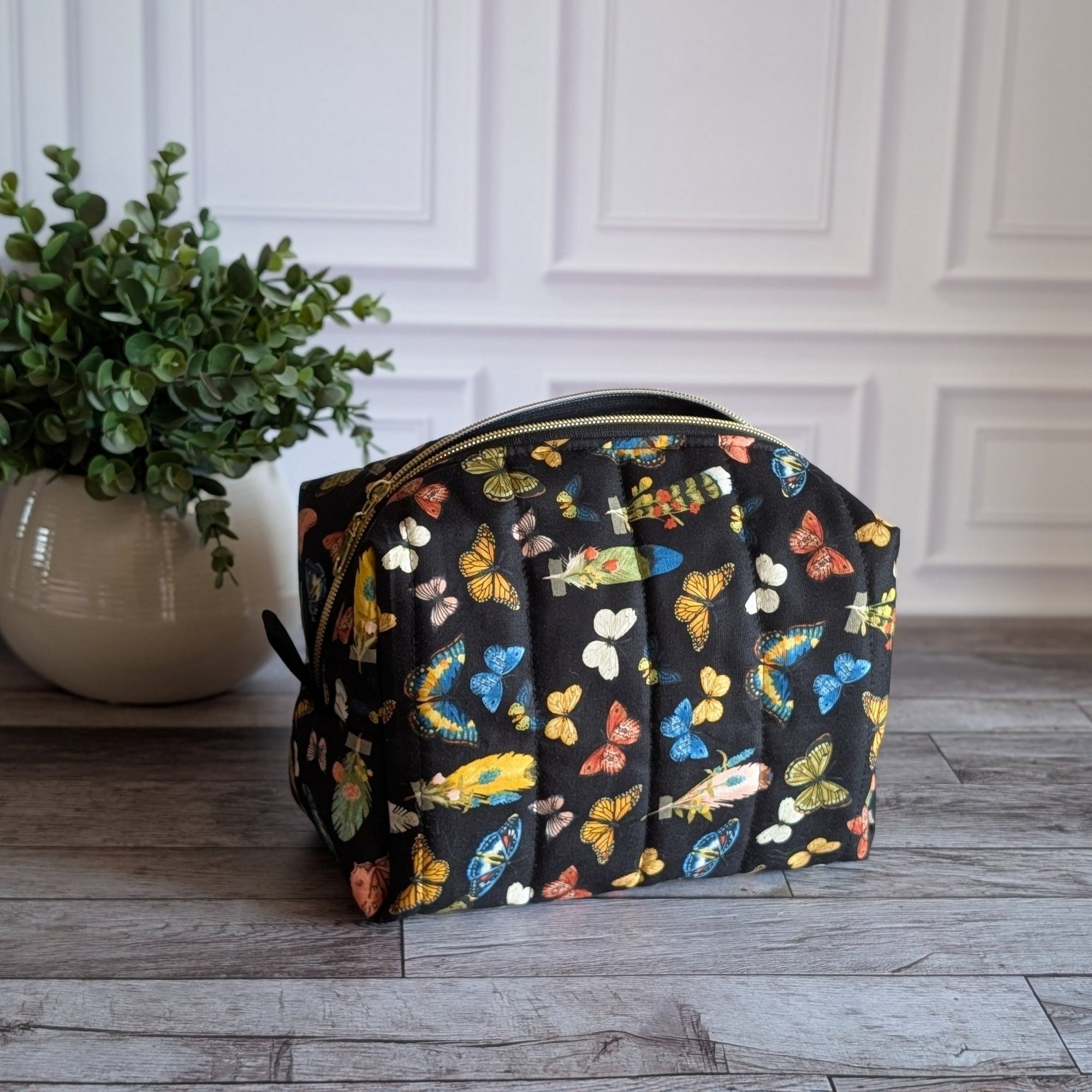 A black zippered cosmetic pouch decorated with a colorful butterfly and heart pattern, set on a gray wood floor next to a potted green plant against a white paneled wall.