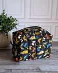 A black zippered cosmetic pouch decorated with a colorful butterfly and heart pattern, set on a gray wood floor next to a potted green plant against a white paneled wall.