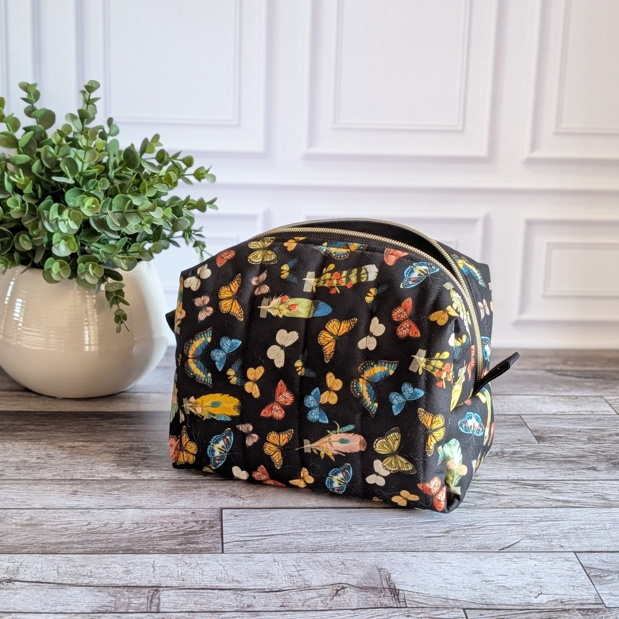 A black zippered cosmetic pouch decorated with a colorful butterfly pattern, set on a gray wood floor next to a potted green plant against a white paneled wall.