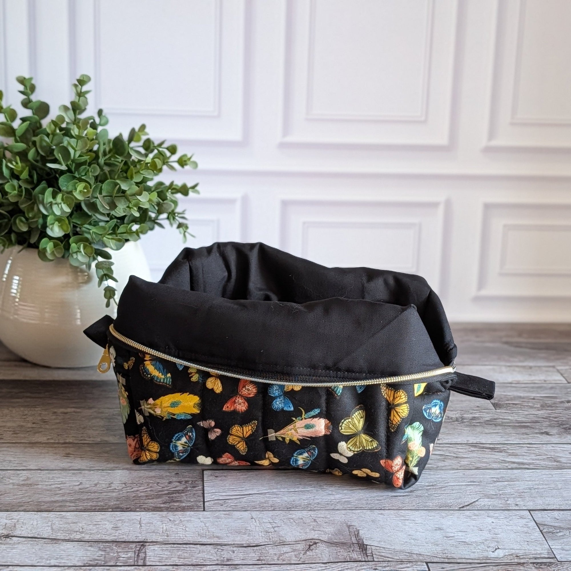 A black zippered cosmetic pouch decorated with a colorful butterfly pattern, set on a gray wood floor next to a potted green plant against a white paneled wall.