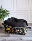 A black zippered cosmetic pouch decorated with a colorful butterfly pattern, set on a gray wood floor next to a potted green plant against a white paneled wall.