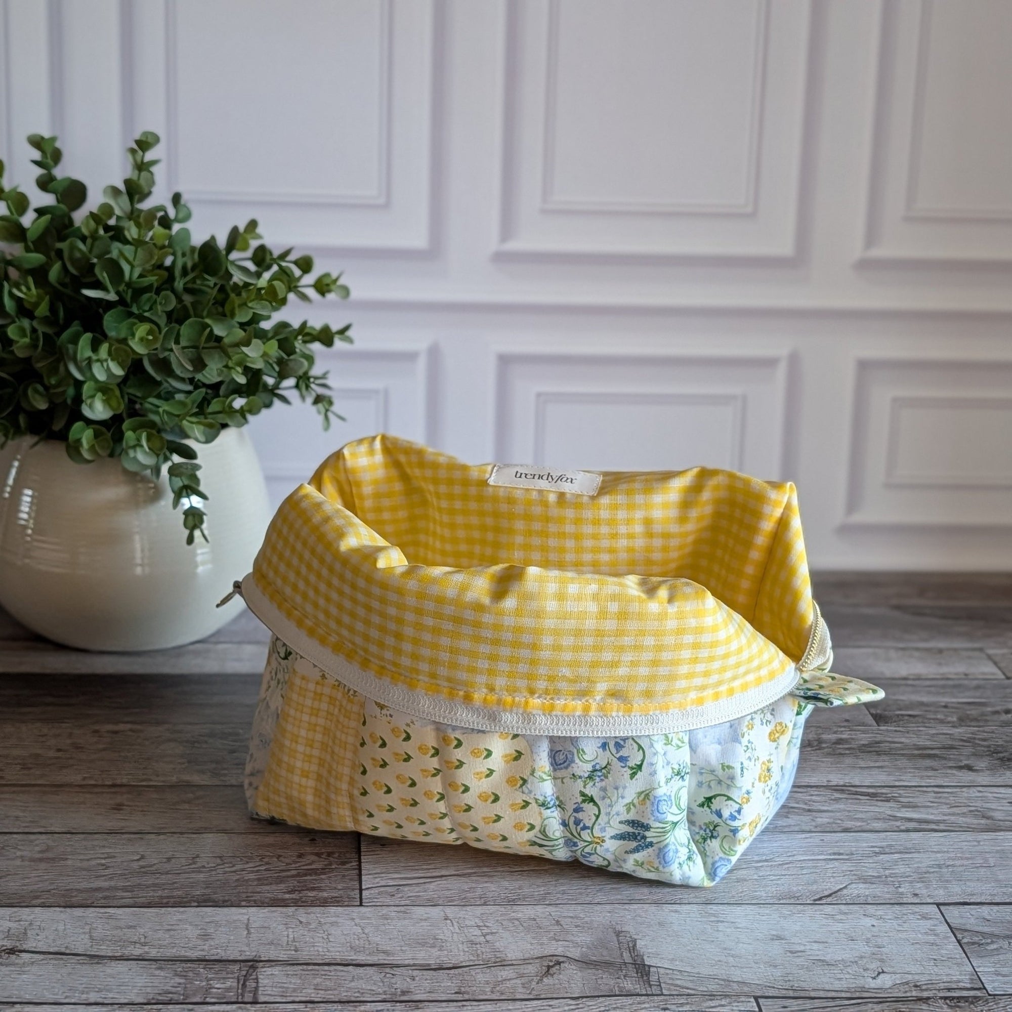 A handmade patchwork cosmetic bag made from yellow, white, and floral fabrics, sitting on a gray wood floor in front of a white paneled wall with a potted plant on the left.