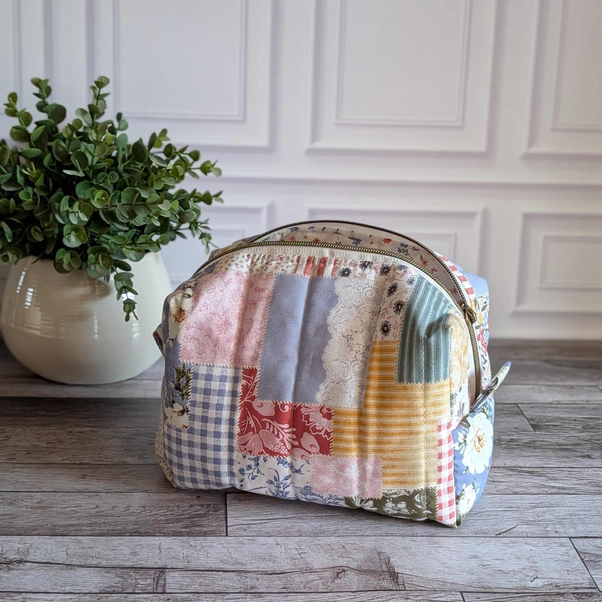 A patchwork fabric cosmetic pouch with a curved top and zip closure, displayed on a gray wood floor next to a potted green plant against a white paneled wall.