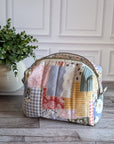 A patchwork fabric cosmetic pouch with a curved top and zip closure, displayed on a gray wood floor next to a potted green plant against a white paneled wall.