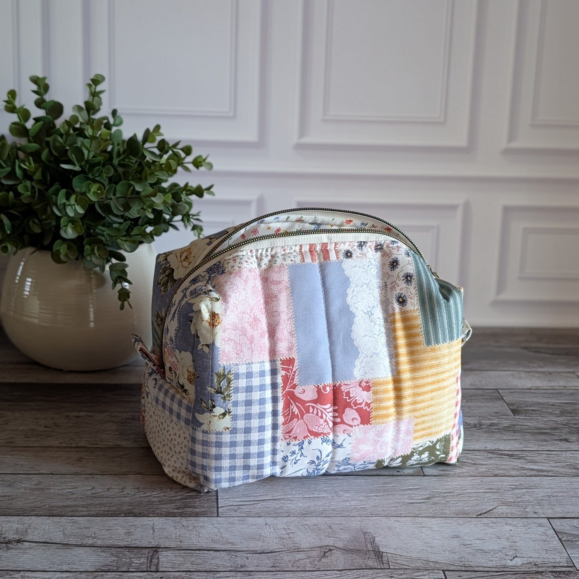 A patchwork fabric cosmetic pouch with a curved top and zip closure, displayed on a gray wood floor next to a potted green plant against a white paneled wall.
