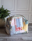 A patchwork fabric cosmetic pouch with a curved top and zip closure, displayed on a gray wood floor next to a potted green plant against a white paneled wall.