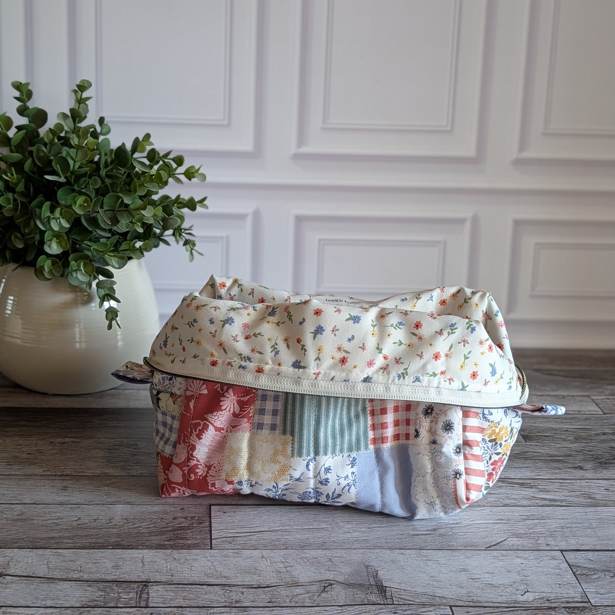 A patchwork fabric cosmetic pouch with a curved top and zip closure, displayed on a gray wood floor next to a potted green plant against a white paneled wall.