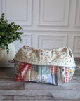 A patchwork fabric cosmetic pouch with a curved top and zip closure, displayed on a gray wood floor next to a potted green plant against a white paneled wall.
