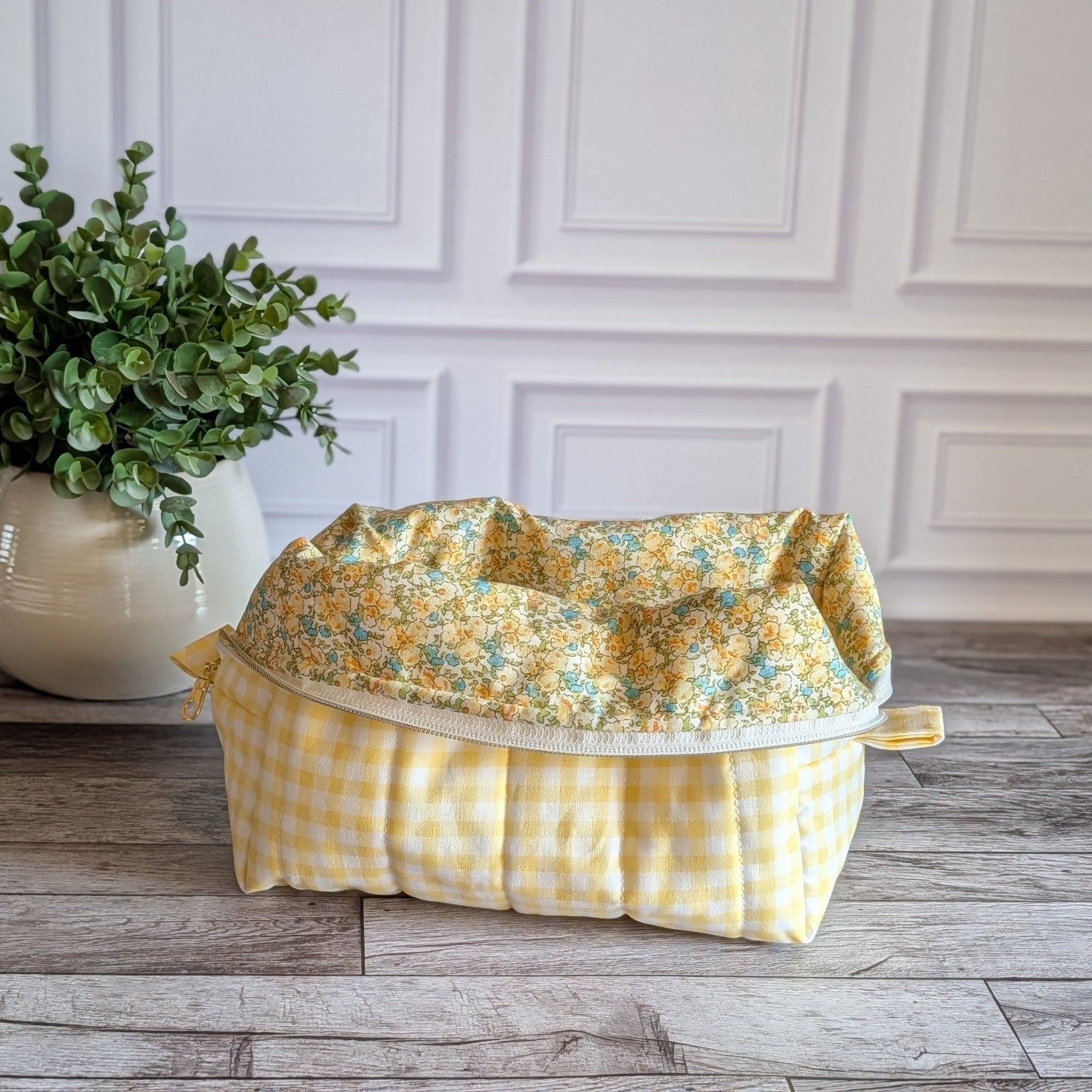 A fabric cosmetic pouch with a yellow and white gingham outer and a yellow floral top lining, placed on a rustic wood floor next to a potted green plant against a white paneled wall.