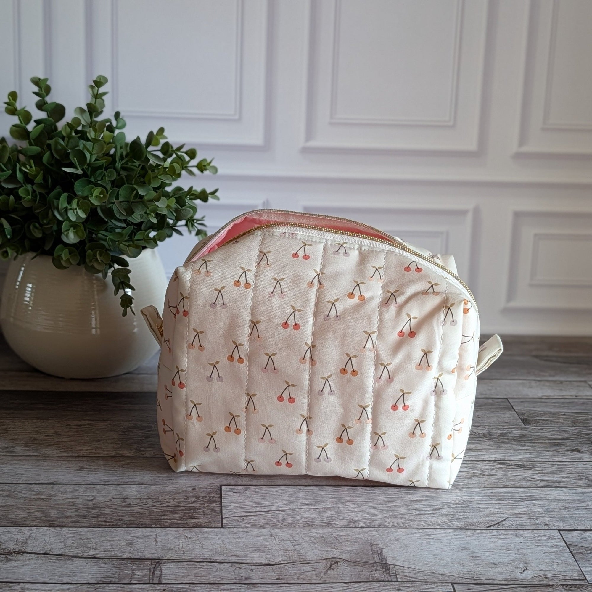A quilted cosmetic pouch with a cream background and a repeating cherry-blossom pattern, set on a gray wood floor next to a potted green plant against a white paneled wall.