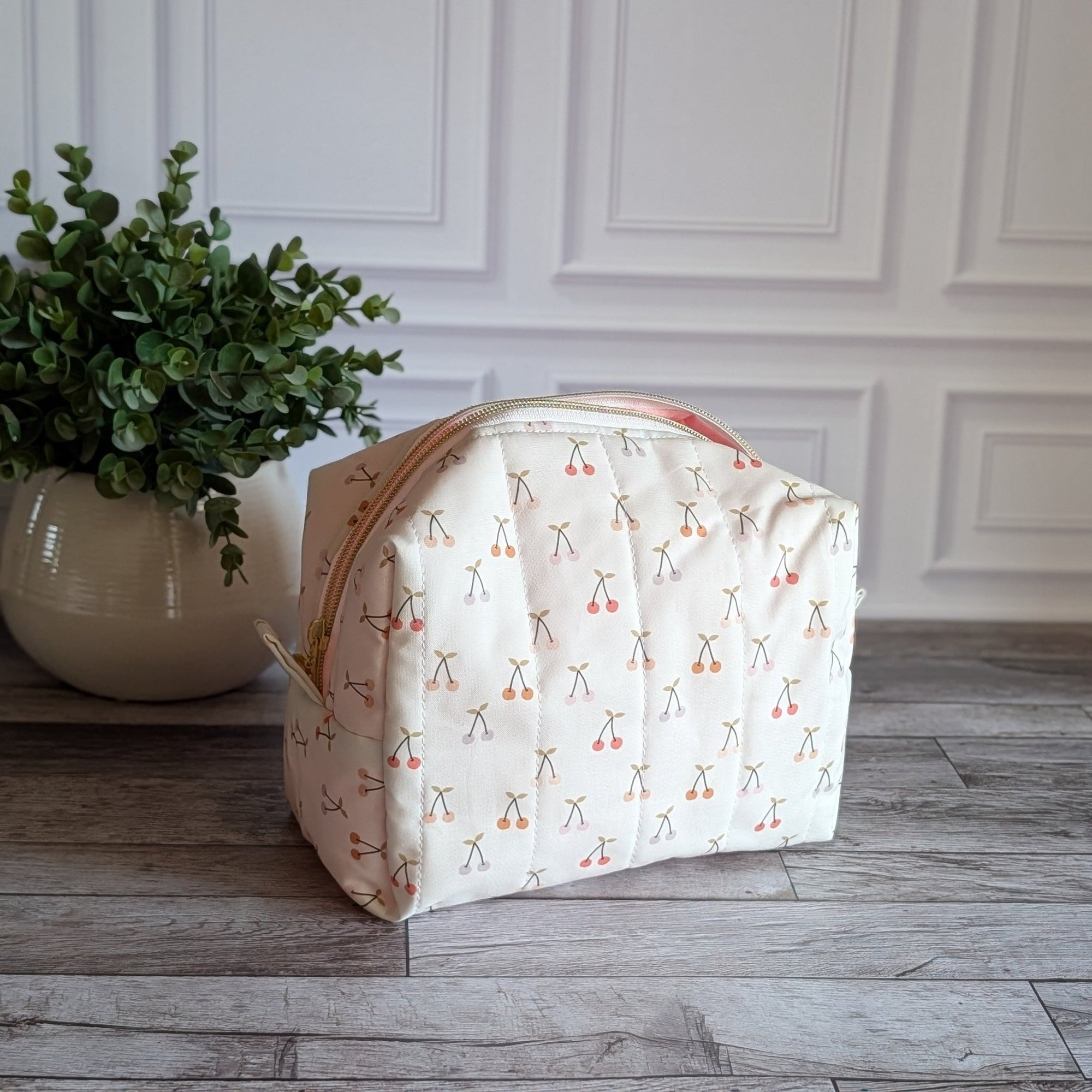 A quilted fabric cosmetic bag with a cherry-and-scales pattern, set on a gray wood floor next to a potted green plant against a white paneled wall.