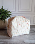 A quilted fabric cosmetic bag with a cherry-and-scales pattern, set on a gray wood floor next to a potted green plant against a white paneled wall.