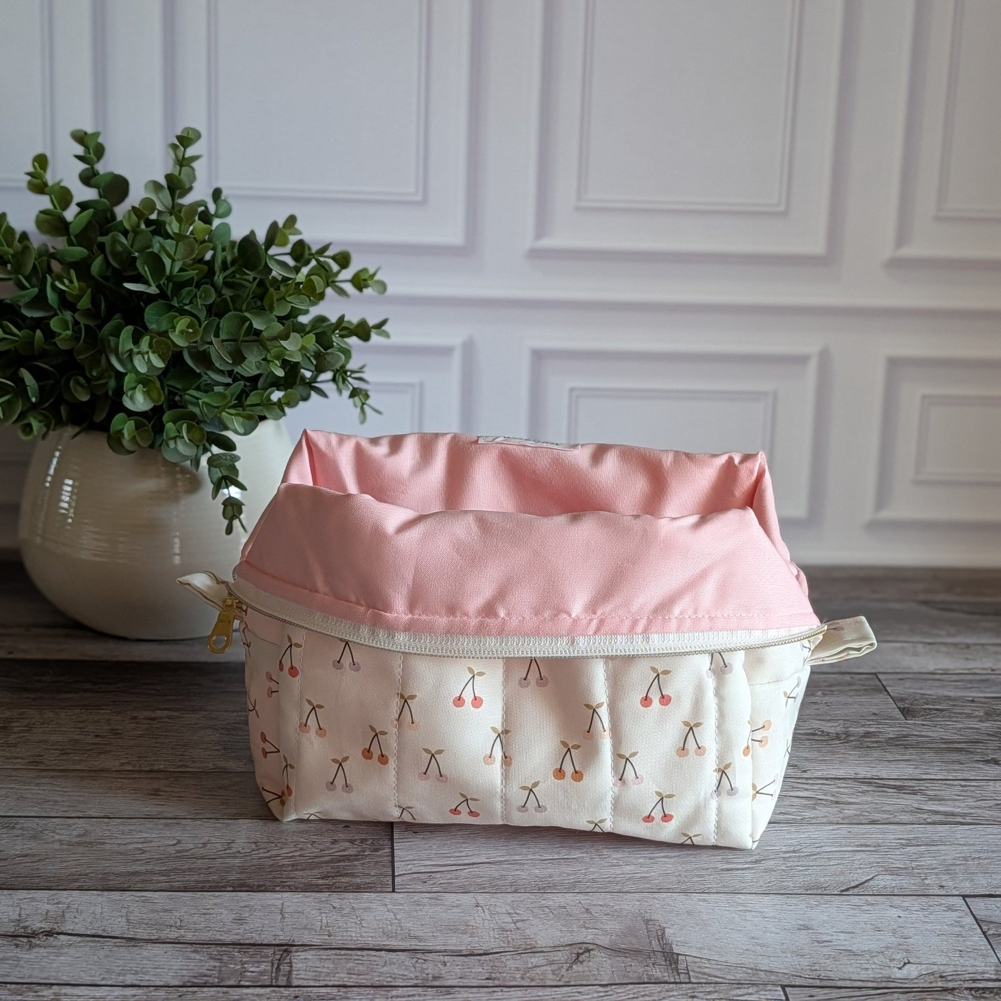 A quilted cosmetic pouch with a cream background and a repeating cherry-blossom pattern, set on a gray wood floor next to a potted green plant against a white paneled wall.