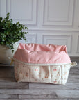 A quilted cosmetic pouch with a cream background and a repeating cherry-blossom pattern, set on a gray wood floor next to a potted green plant against a white paneled wall.