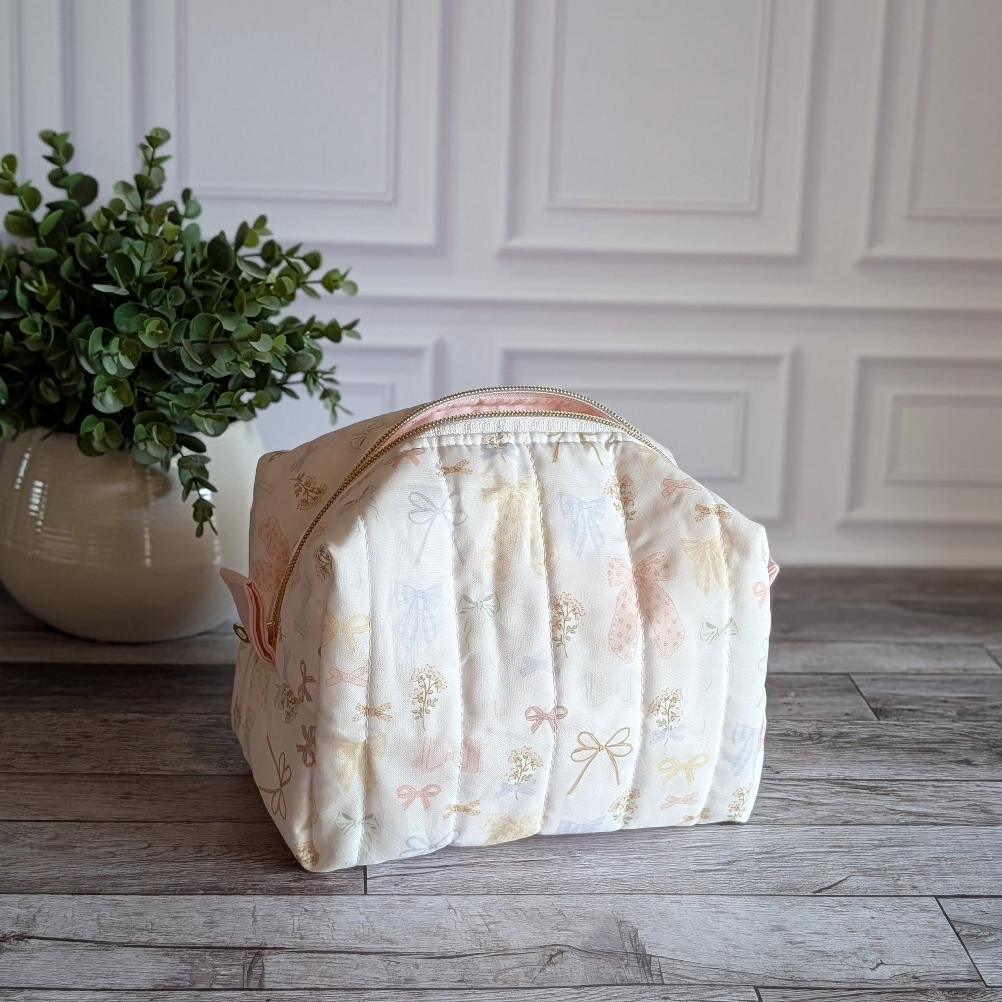 A quilted travel toiletry bag with a cream and pale pink floral pattern sits on a gray wood floor in front of a white paneled wall, next to a potted green plant.