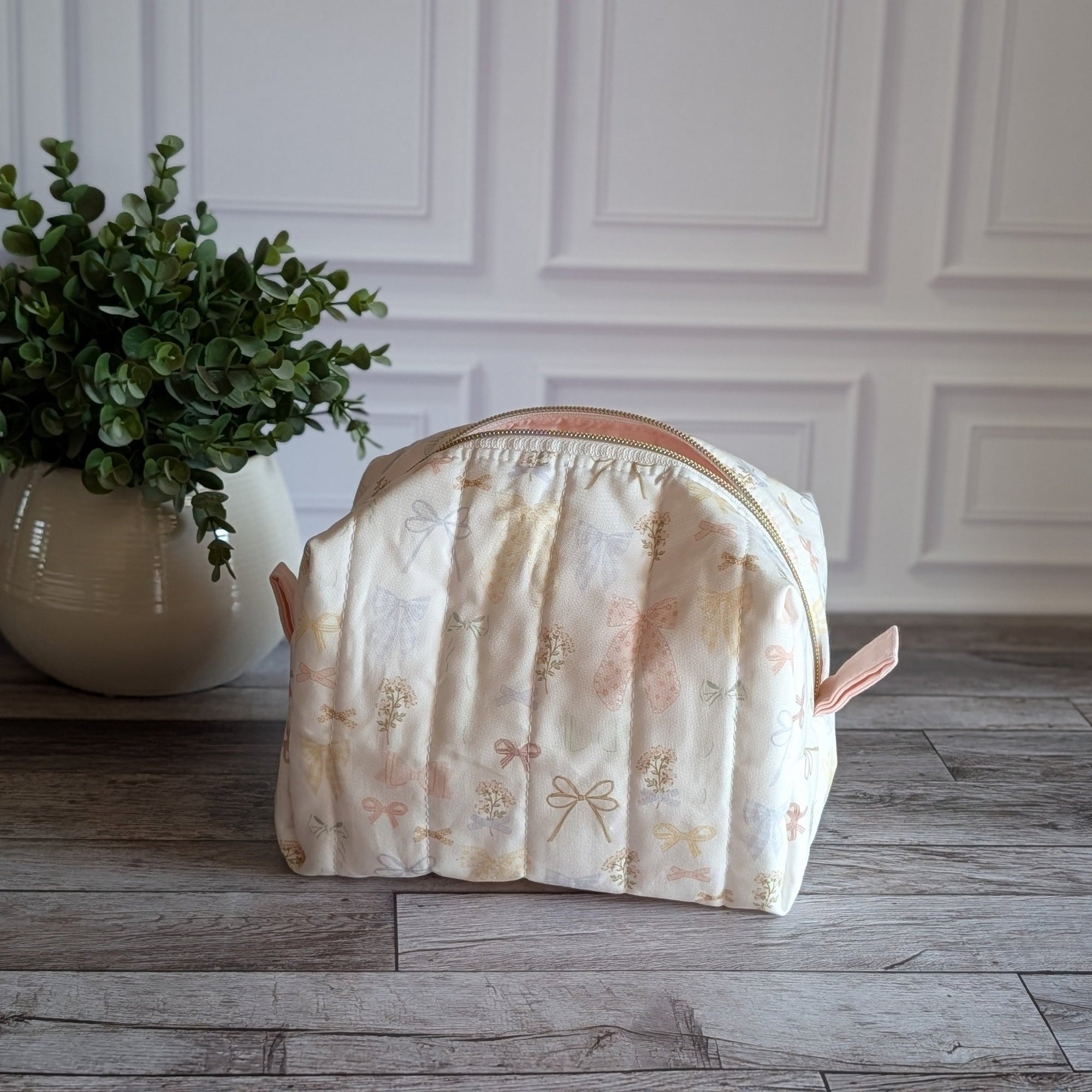 A quilted travel toiletry bag with a cream and pale pink floral pattern sits on a gray wood floor in front of a white paneled wall, next to a potted green plant.