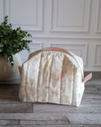 A quilted travel toiletry bag with a cream and pale pink floral pattern sits on a gray wood floor in front of a white paneled wall, next to a potted green plant.