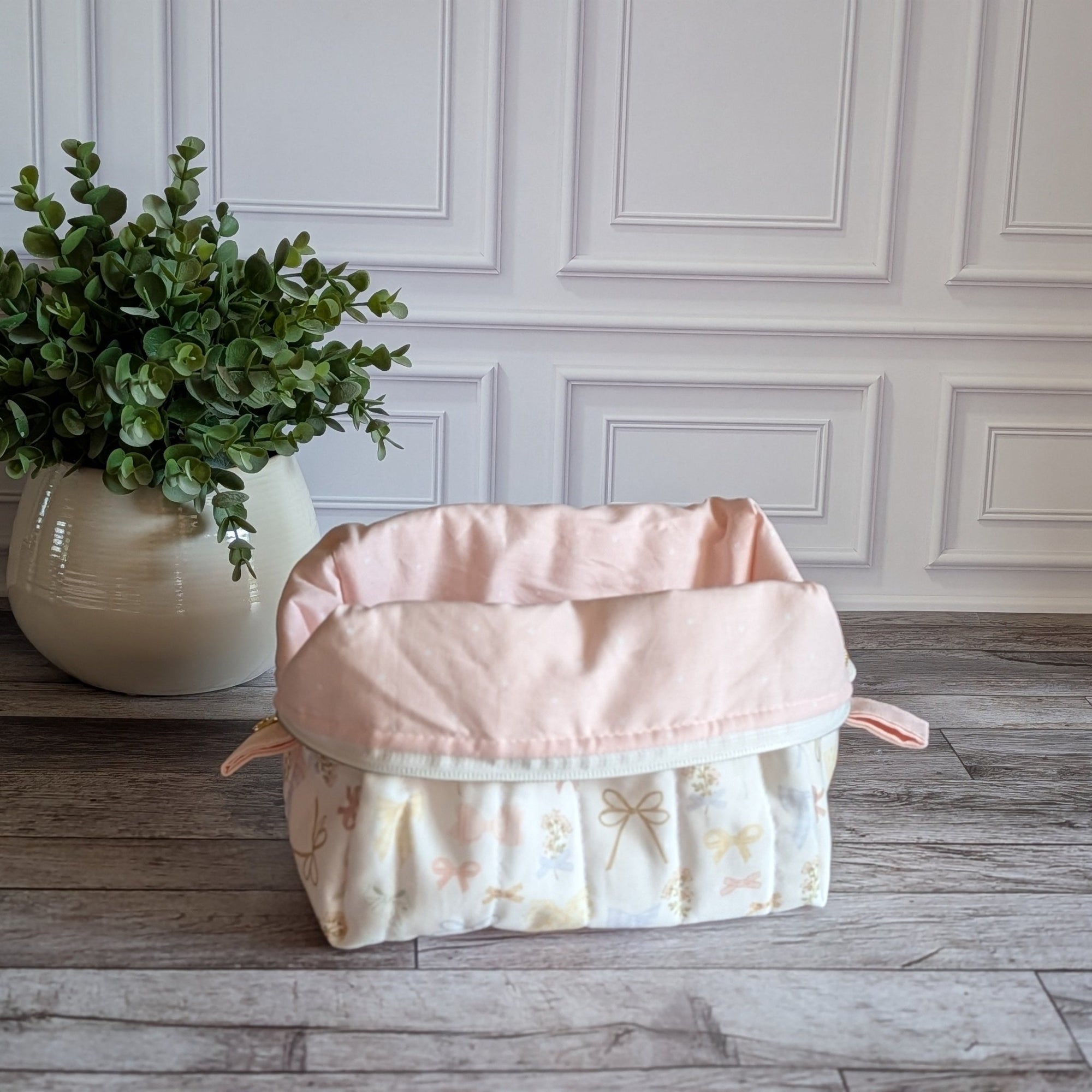 A quilted travel toiletry bag with a cream and pale pink floral pattern sits on a gray wood floor in front of a white paneled wall, next to a potted green plant.
