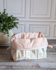 A quilted travel toiletry bag with a cream and pale pink floral pattern sits on a gray wood floor in front of a white paneled wall, next to a potted green plant.