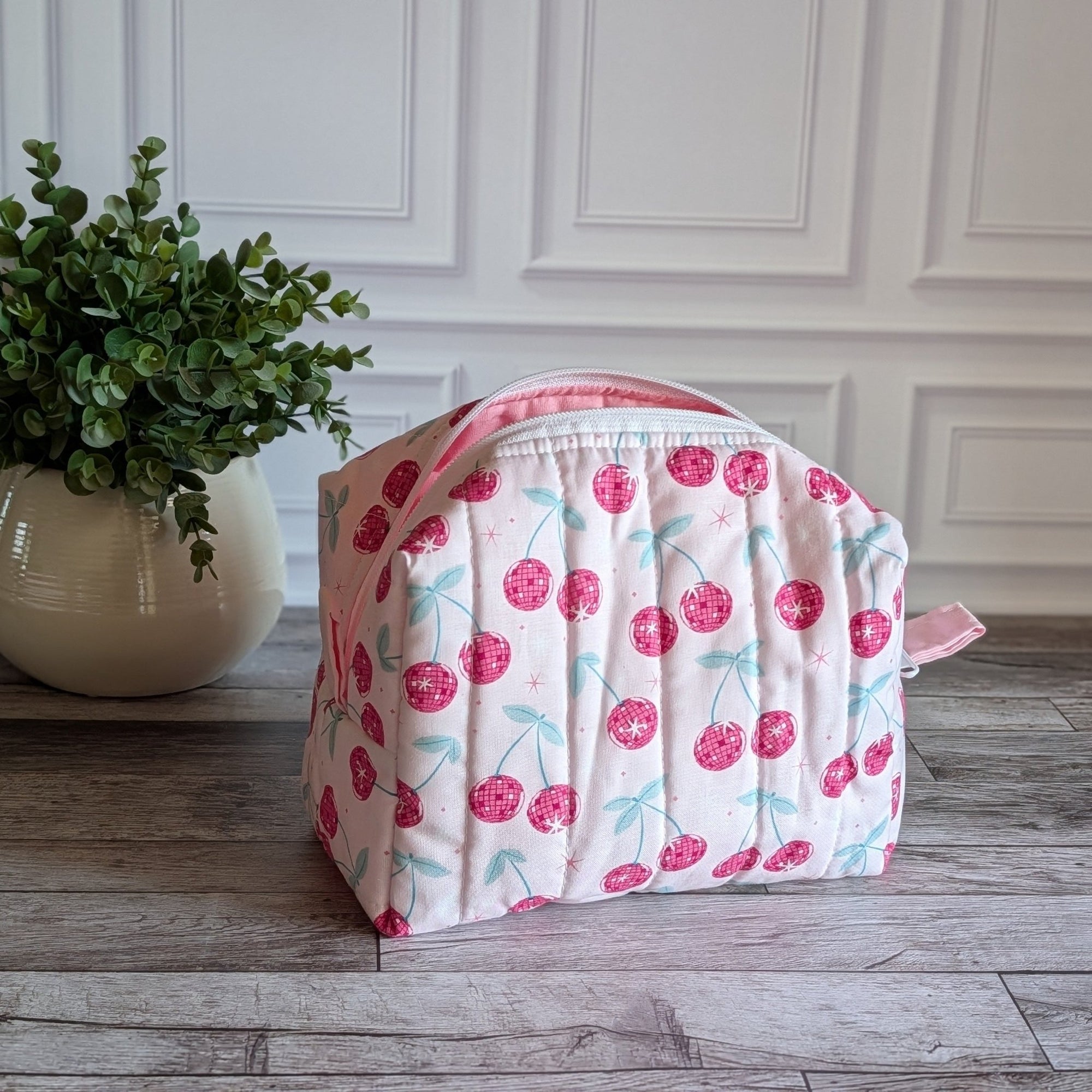 A quilted cosmetic bag with a pink and white cherry pattern, placed on a gray wood floor next to a potted green plant against a white paneled wall.