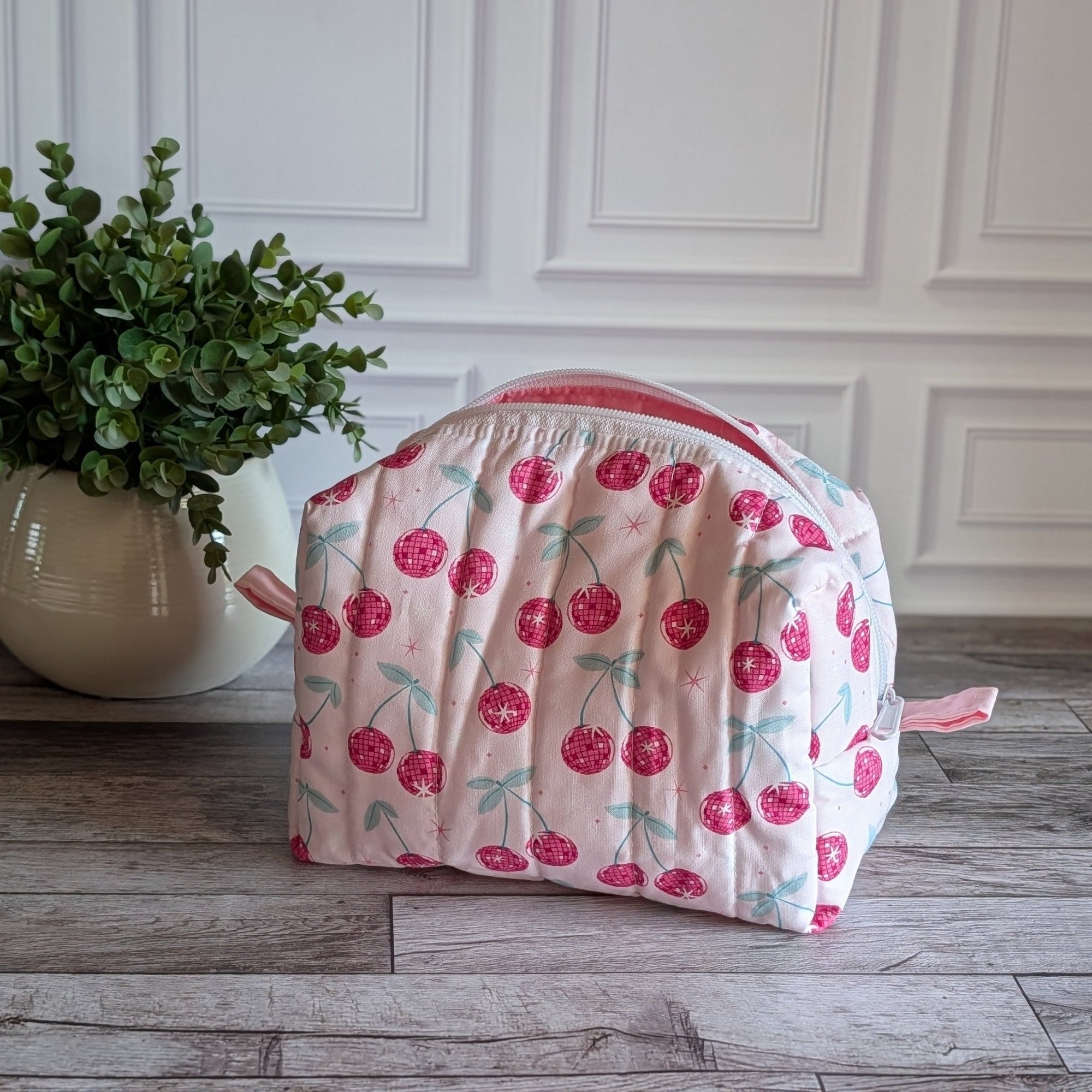 A quilted cosmetic bag with a pink and white cherry pattern, placed on a gray wood floor next to a potted green plant against a white paneled wall.