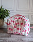 A quilted cosmetic bag with a pink and white cherry pattern, placed on a gray wood floor next to a potted green plant against a white paneled wall.