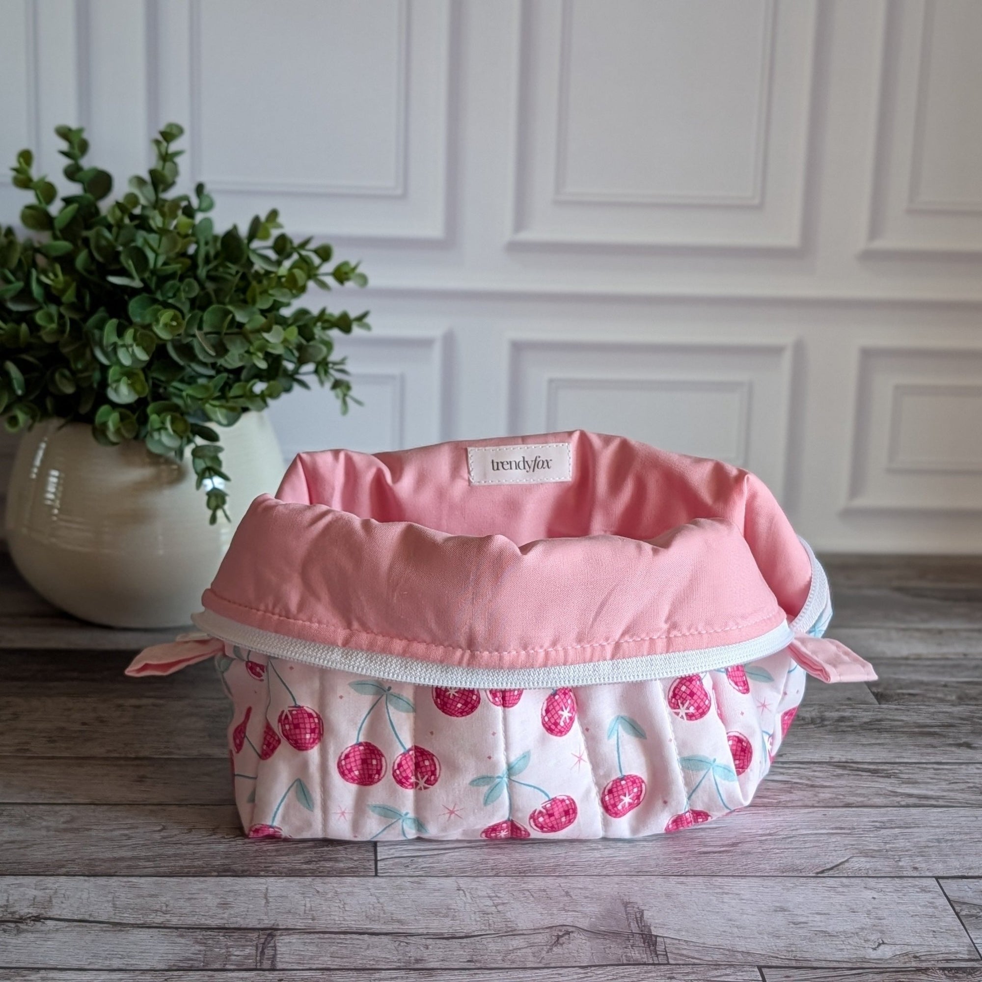 A quilted cosmetic bag with a pink and white cherry pattern, placed on a gray wood floor next to a potted green plant against a white paneled wall.