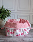 A quilted cosmetic bag with a pink and white cherry pattern, placed on a gray wood floor next to a potted green plant against a white paneled wall.