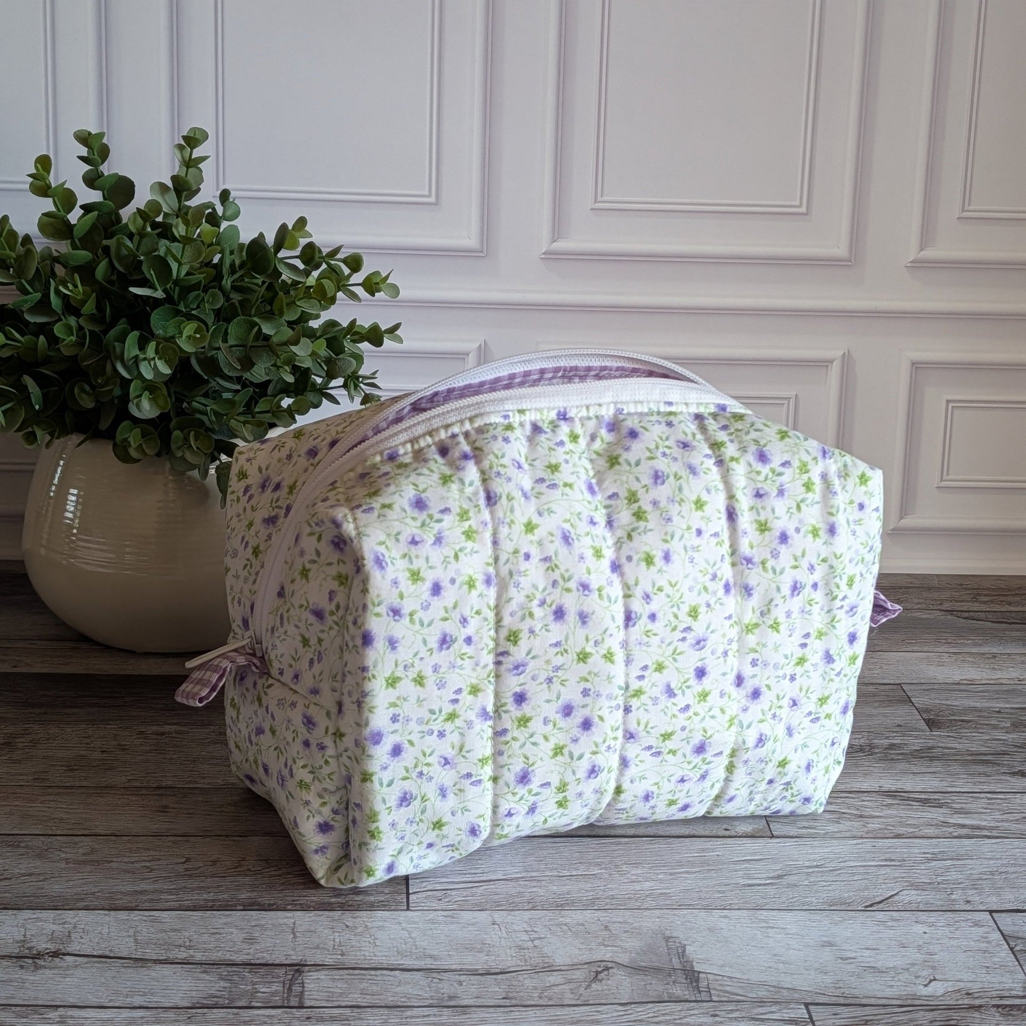 A quilted cosmetic bag with a white background and a purple and green floral pattern, placed on a gray wood floor next to a potted green plant.
