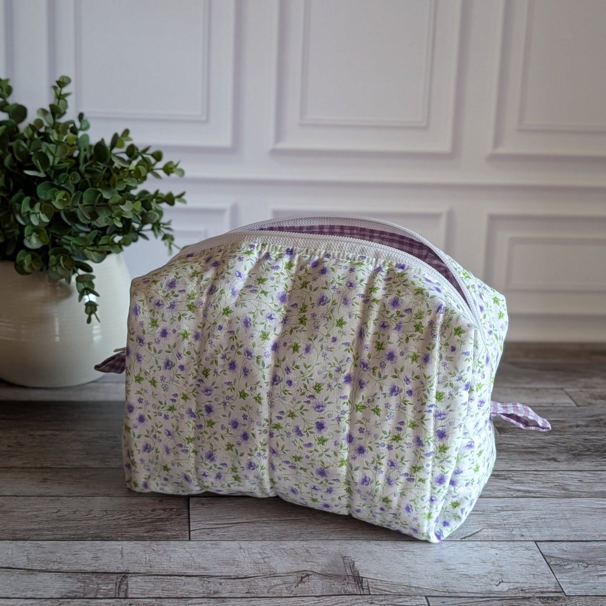 A quilted cosmetic bag with a white, purple and green floral pattern and purple gingham lining, placed on a gray wood floor next to a potted green plant.