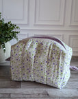 A quilted cosmetic bag with a white, purple and green floral pattern and purple gingham lining, placed on a gray wood floor next to a potted green plant.