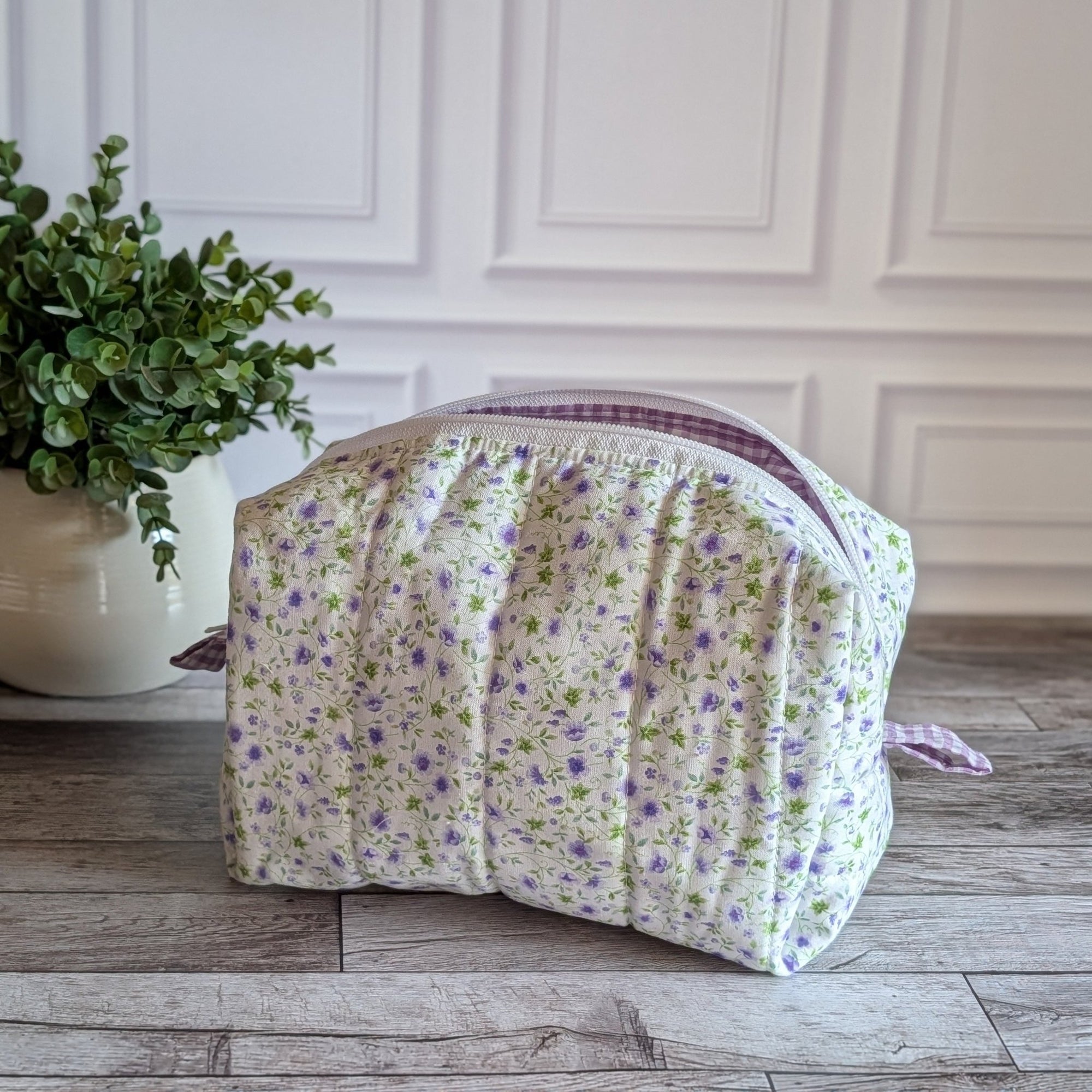 A quilted cosmetic bag with a white, purple and green floral pattern and purple gingham lining, placed on a gray wood floor next to a potted green plant.