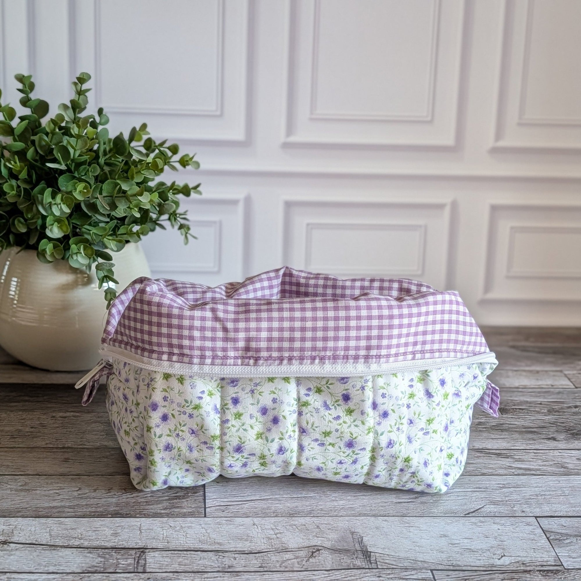 A quilted cosmetic bag with a white, purple and green floral pattern and purple gingham lining, placed on a gray wood floor next to a potted green plant.