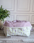 A quilted cosmetic bag with a white, purple and green floral pattern and purple gingham lining, placed on a gray wood floor next to a potted green plant.