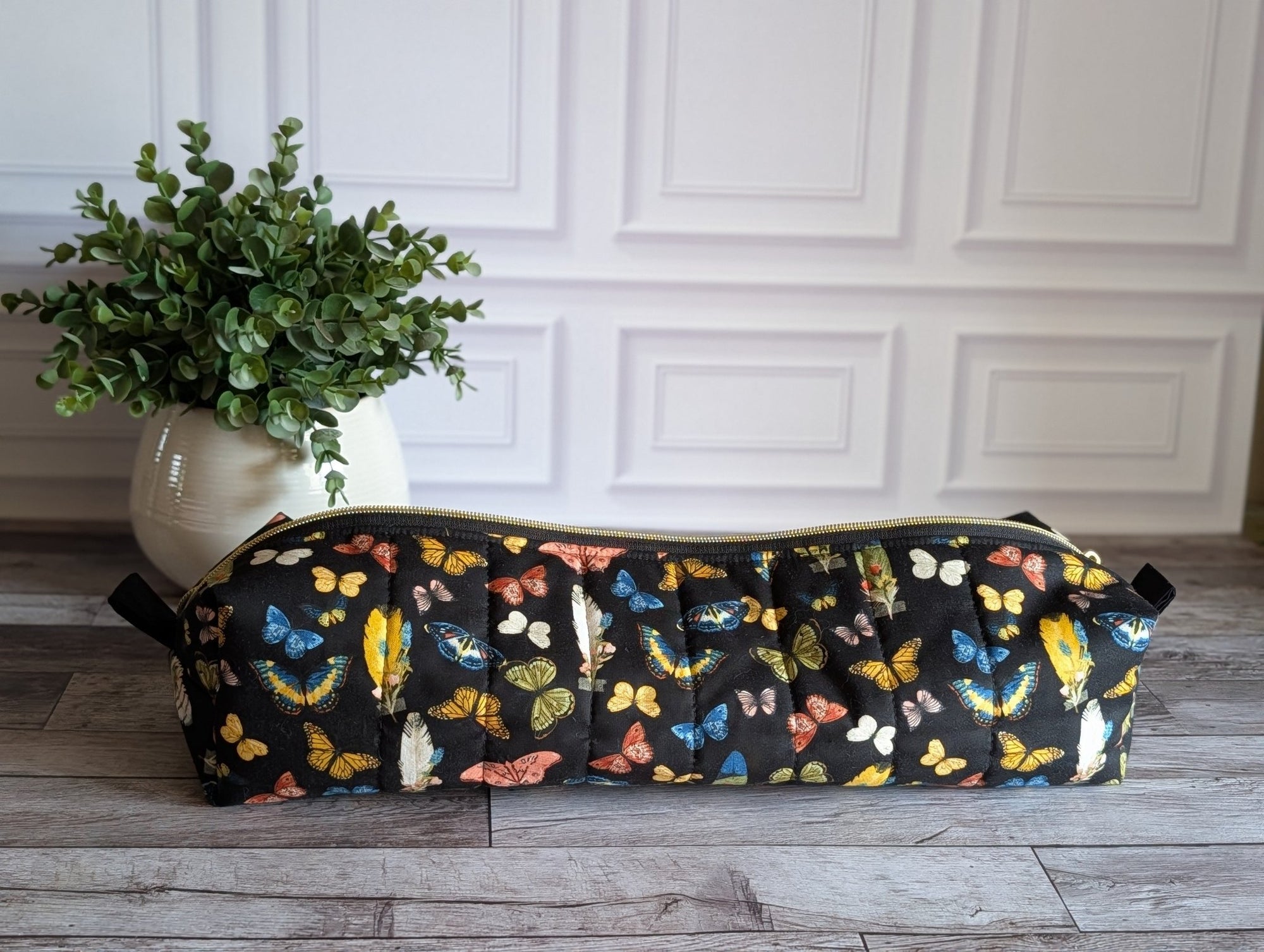 A long cylindrical black zipper pouch with a colorful butterfly print, resting on a gray wood floor next to a potted green plant against a white paneled wall.