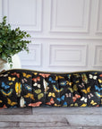 A long cylindrical black zipper pouch with a colorful butterfly print, resting on a gray wood floor next to a potted green plant against a white paneled wall.