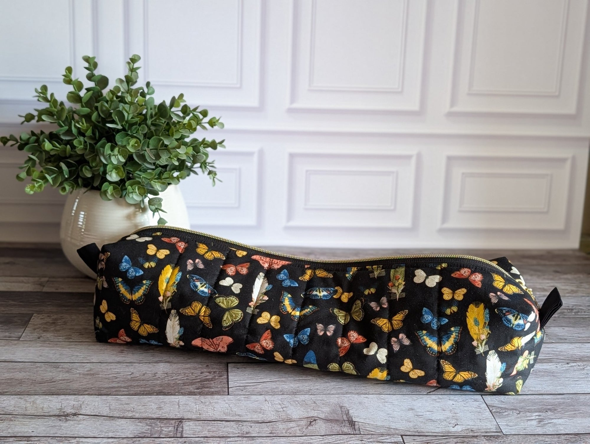 A long cylindrical black zipper pouch with a colorful butterfly print, resting on a gray wood floor next to a potted green plant against a white paneled wall.