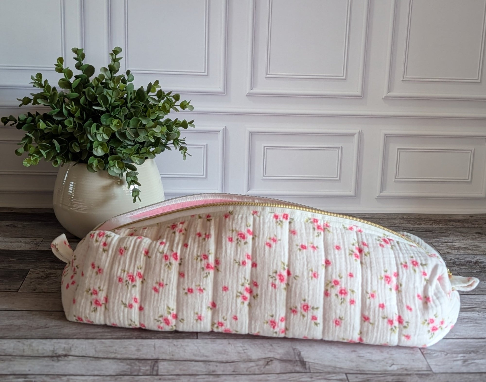 A quilted fabric toiletry bag with a pink floral print and a pink zipper, resting on a gray wooden floor next to a potted green plant against a white paneled wall.