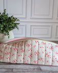 A quilted fabric toiletry bag with a pink floral print and a pink zipper, resting on a gray wooden floor next to a potted green plant against a white paneled wall.
