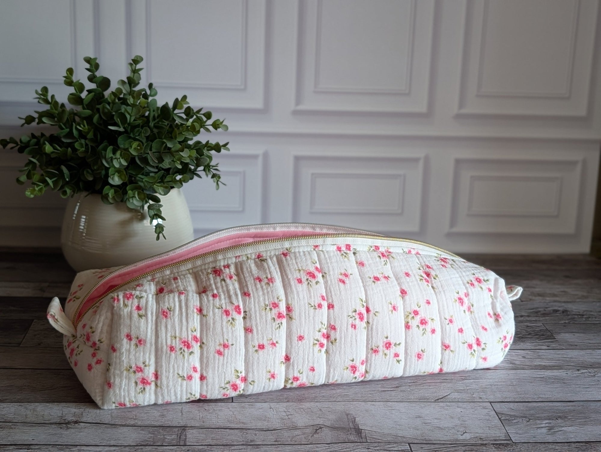 A quilted fabric makeup bag with a pink floral print and a pink zipper, resting on a gray wooden floor next to a potted green plant against a white paneled wall.