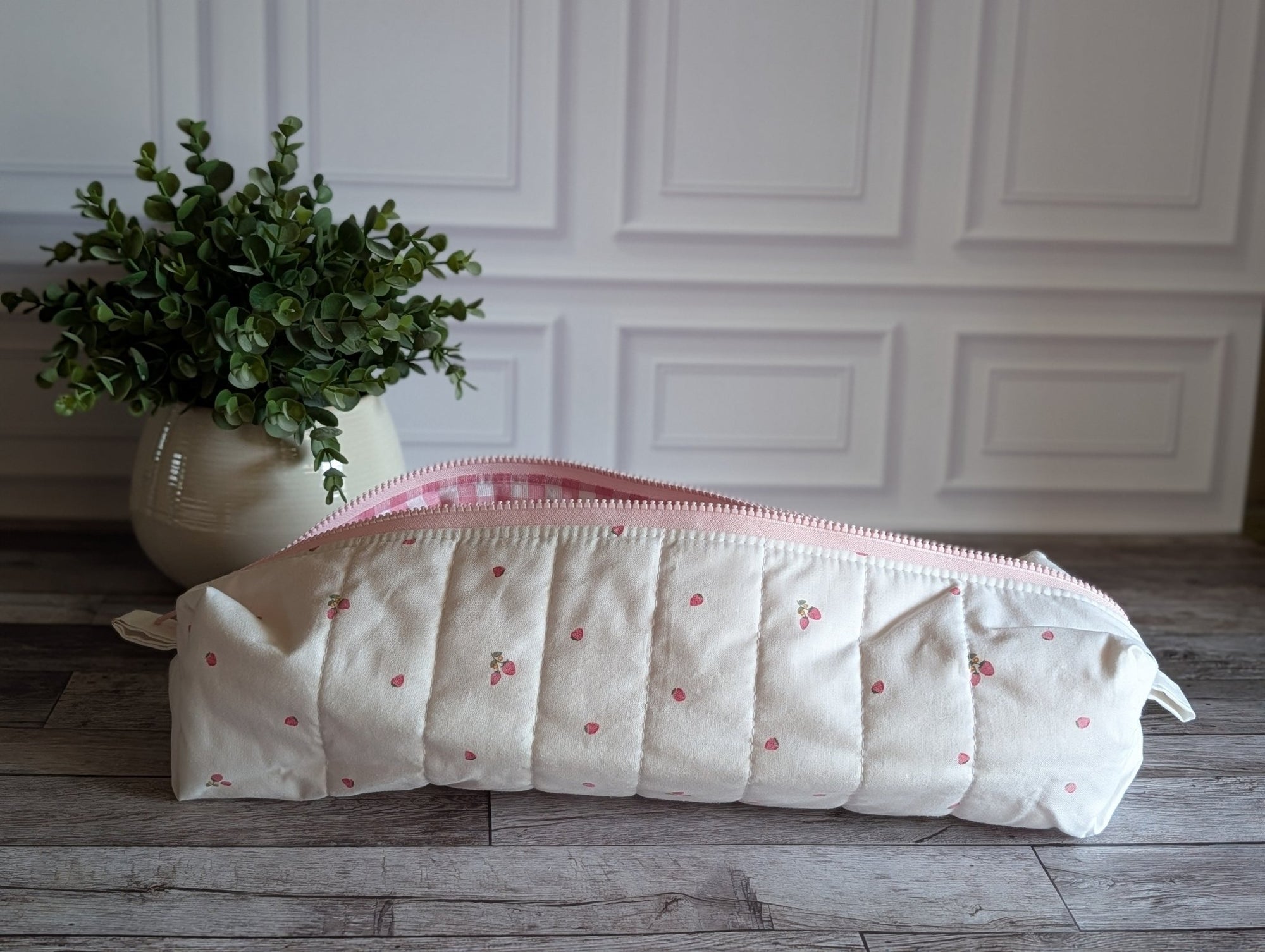 A quilted cream-colored pencil case with a pink cherry pattern and a pink gingham lining, resting on a gray wooden floor next to a potted green plant.