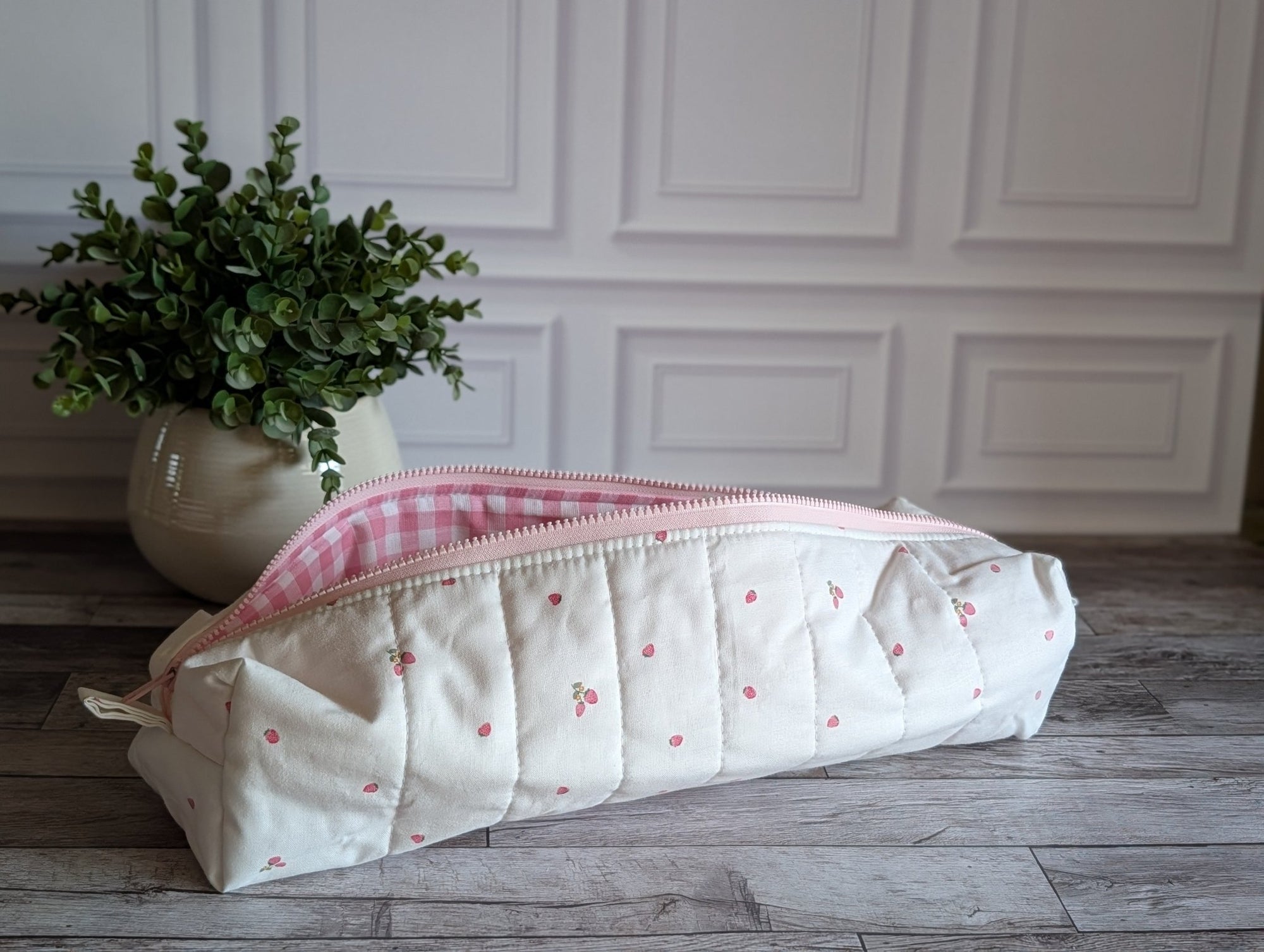 A quilted fabric zippered pouch with a cream background, small pink cherry pattern, and a pink gingham lining, resting on a gray wood floor next to a potted green plant.
