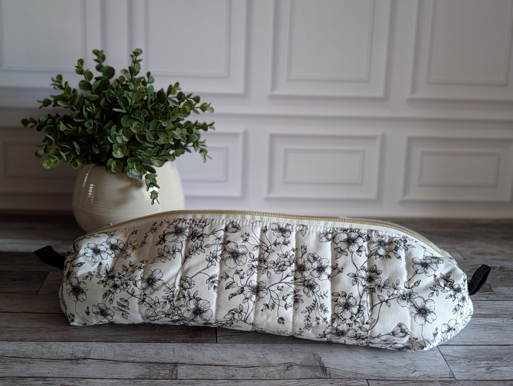 A quilted hair tool pouch with a black floral print on a white background, resting on a gray wooden floor in front of a white paneled wall.