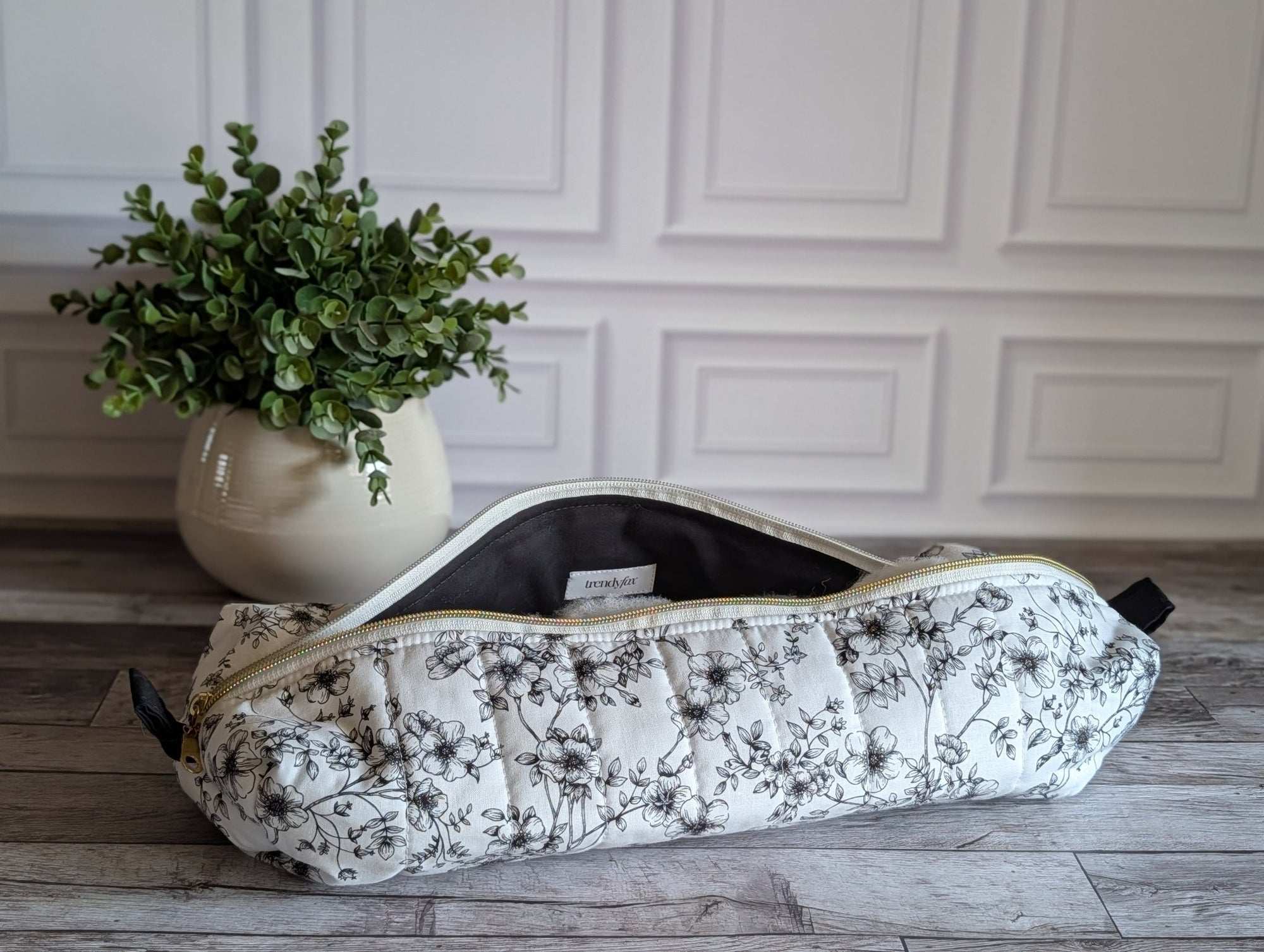 A quilted hair tool pouch with a black floral print on a white background, resting on a gray wooden floor in front of a white paneled wall.