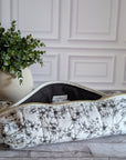 A quilted hair tool pouch with a black floral print on a white background, resting on a gray wooden floor in front of a white paneled wall.